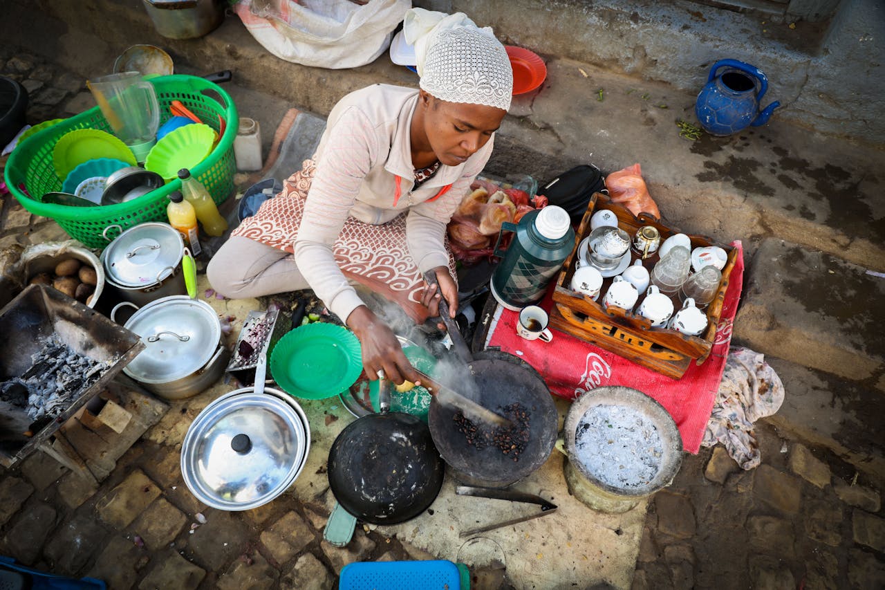 A woman in a white knit cap and light-colored clothing prepares traditional Kenyan coffee in a rustic outdoor setting. She crouches beside various metal pots and pans arranged on the ground, with a charcoal fire visible to the left. Ceramic cups, bottles, and supplies are organized on a red cloth. The scene captures the authentic, hands-on craft of traditional coffee preparation, conveying warmth, cultural heritage, and community-centered hospitality.